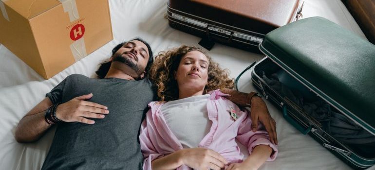 a woman and man lying tired on the bed full of suitcases and boxes