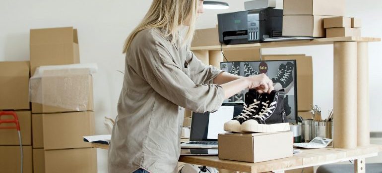 Blond woman packing converse shoes on a home office desk full of boxes and office supplies. 
