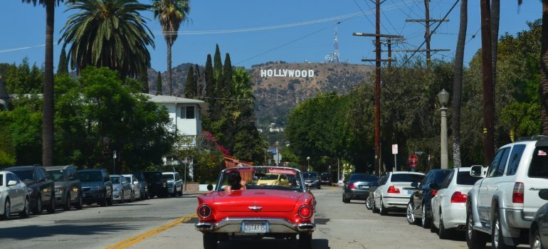 a red car on the road with the Hollywood sign at the top of the hill on the horizon
